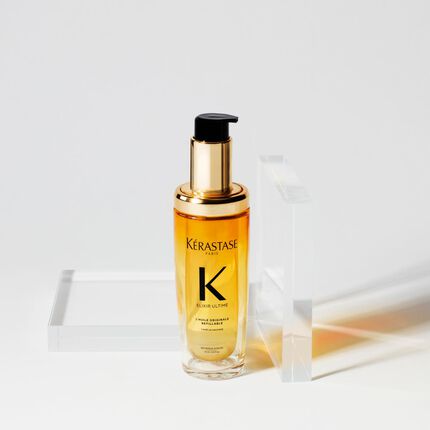 A bottle of Kerastase Elixir Ultime hair oil with a gold pump cap, placed beside a clear rectangular container on a light background.