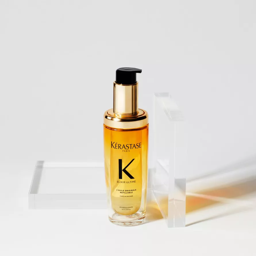 A bottle of Kerastase Elixir Ultime hair oil with a gold pump cap, placed beside a clear rectangular container on a light background.