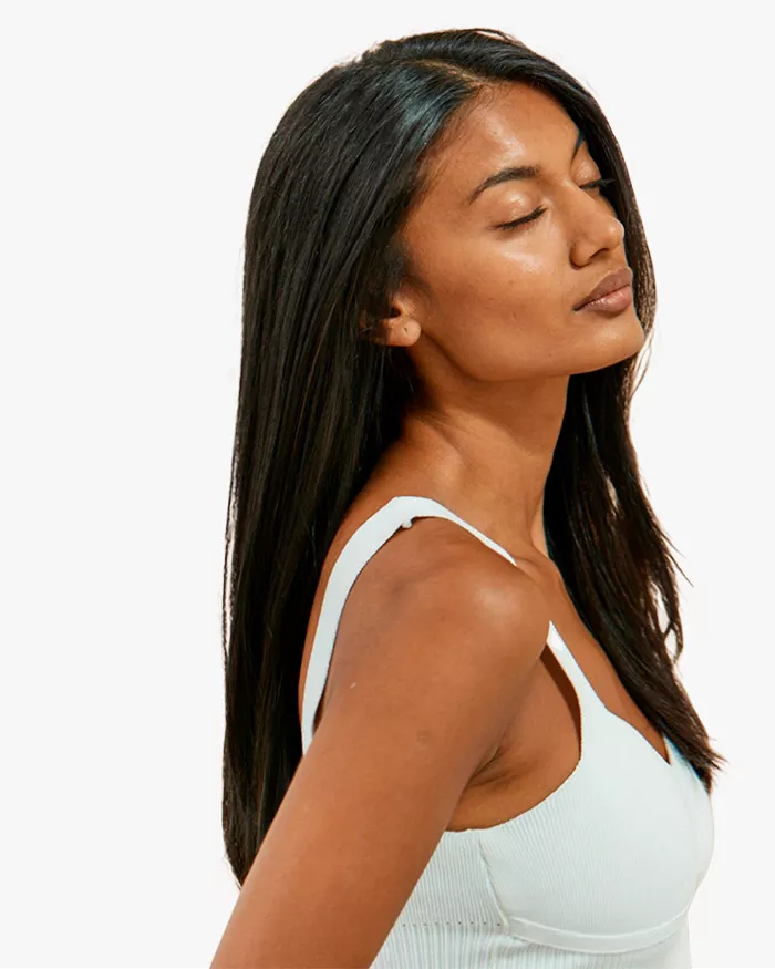 A young woman with long, straight hair is gazing upwards with her eyes closed, wearing a light blue tank top.