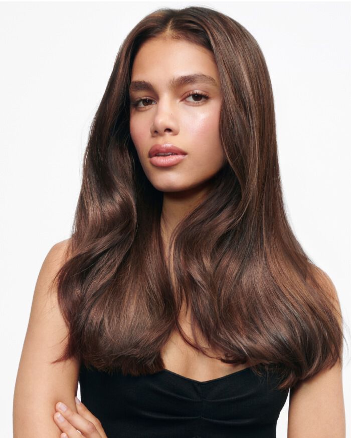 A young person with long, wavy brown hair and natural makeup poses confidently, wearing a black top against a plain white background.