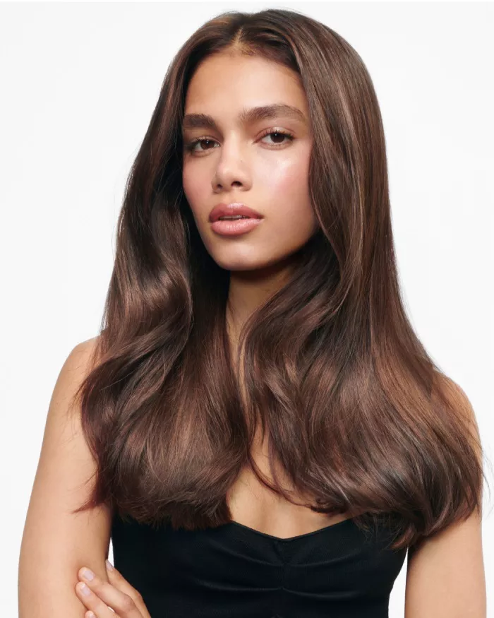 A young person with long, wavy brown hair and natural makeup poses confidently, wearing a black top against a plain white background.