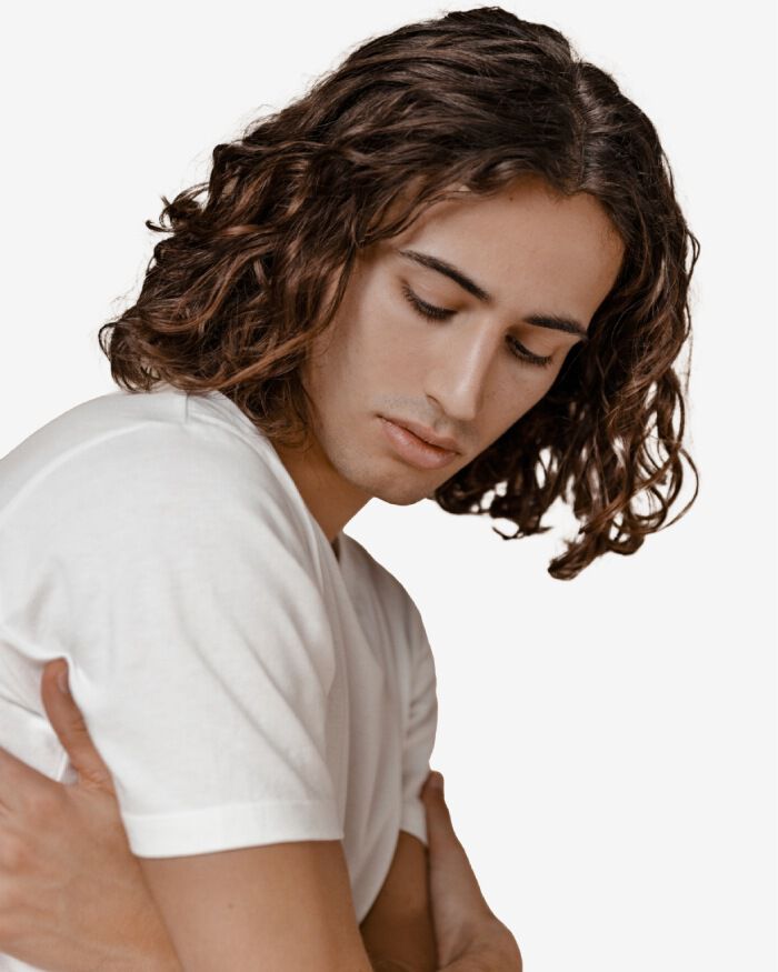 A young man with long, curly hair is looking down thoughtfully while hugging himself, wearing a plain white t-shirt. The background is a soft, light color.