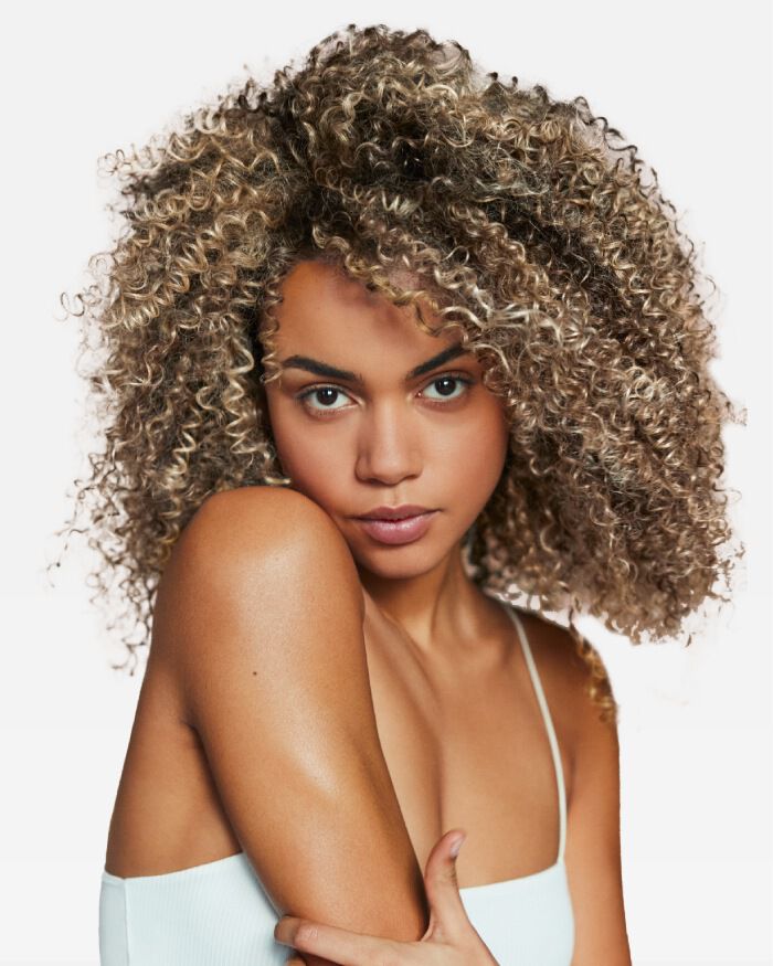 A close-up portrait of a young woman with voluminous, curly hair. She is looking directly at the camera with a soft expression, her arm resting on her shoulder. The background is plain and light-colored.