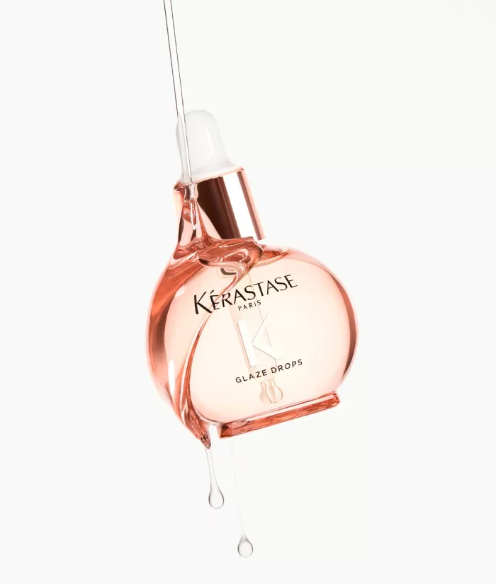 A bottle of Kerastase Glaze Drops serum, round and pink, with a dropper, with a drop of liquid dripping from the tip. The background is white, highlighting the product.