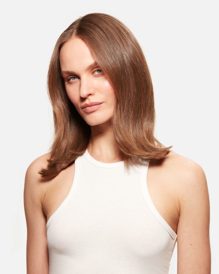 A person with long, straight hair and a neutral expression is looking directly at the camera. They are wearing a simple white sleeveless top against a plain background.