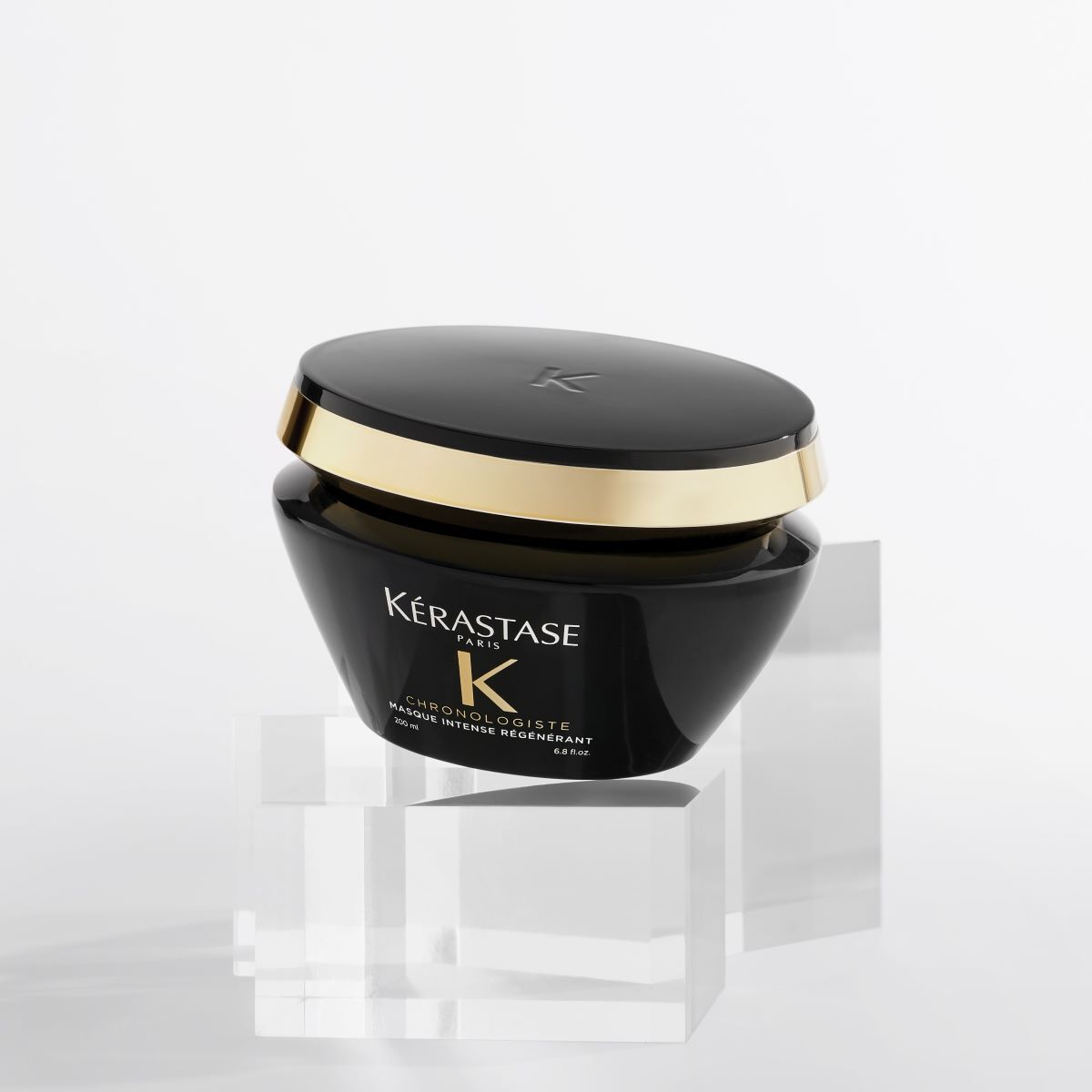 A luxurious black hair treatment mask from Kerastase, featuring a gold accent on the lid, displayed on clear acrylic stands against a minimalist background.