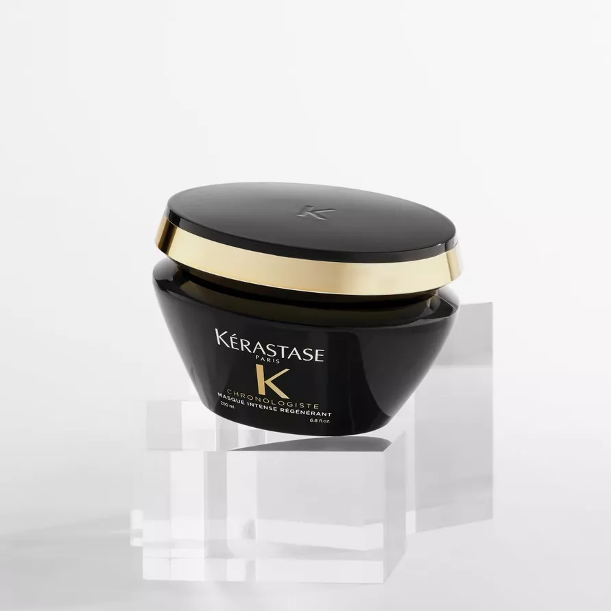 A luxurious black hair treatment mask from Kerastase, featuring a gold accent on the lid, displayed on clear acrylic stands against a minimalist background.