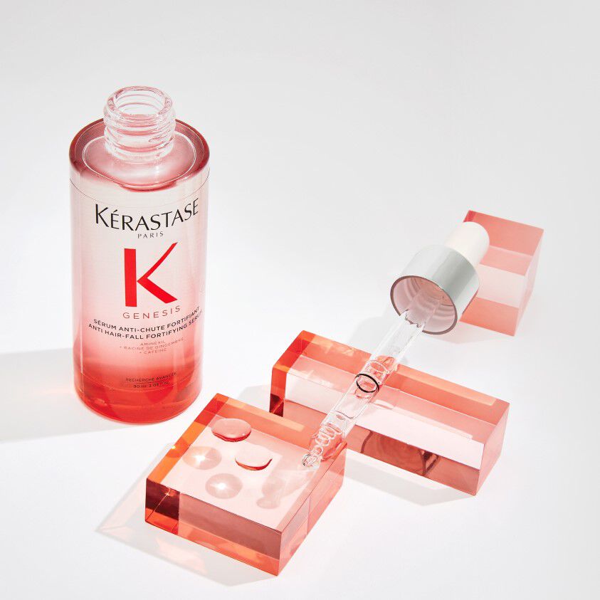 A bottle of Kerastase Genesis serum, featuring a dropper, placed on geometric pink-colored blocks. The label prominently displays the brand name and product line.
