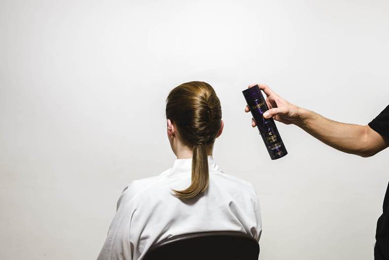 A person with a sleek ponytail is having hair spray applied from behind against a plain white background.