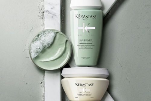 A woman with long, dark hair holds a Kerastase hair product, gazing softly at the camera against a plain background.
