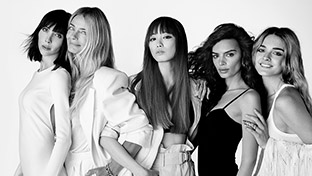 A group of five women, posing closely together in stylish attire, against a simple white background in black and white.