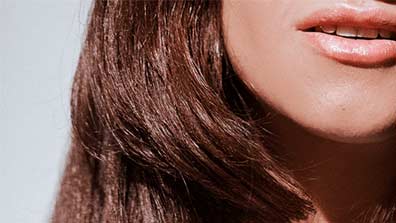 Close-up of a woman's lips and shoulder-length, shiny brown hair against a light background.