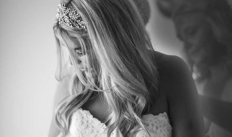 A bride adjusts her dress, wearing an ornate hairpiece, with a soft, reflective background.