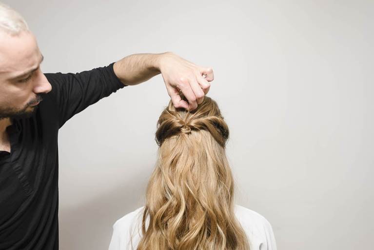 A hairstylist creates an intricate updo on a woman's long, wavy hair, showcasing detailed handiwork against a plain background.