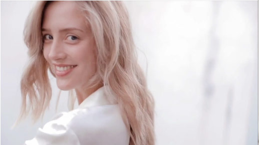 A smiling woman with long, wavy blonde hair poses warmly against a softly blurred white background.