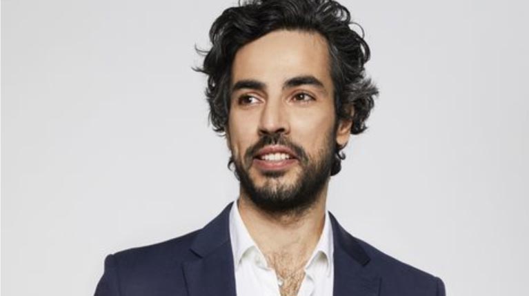 A man with curly hair wearing a dark blazer and white shirt, smiling against a neutral background.