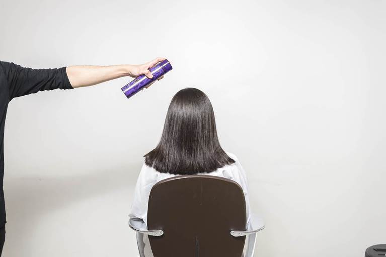 A person sitting in a salon chair has their straight hair styled with hairspray being applied from behind.