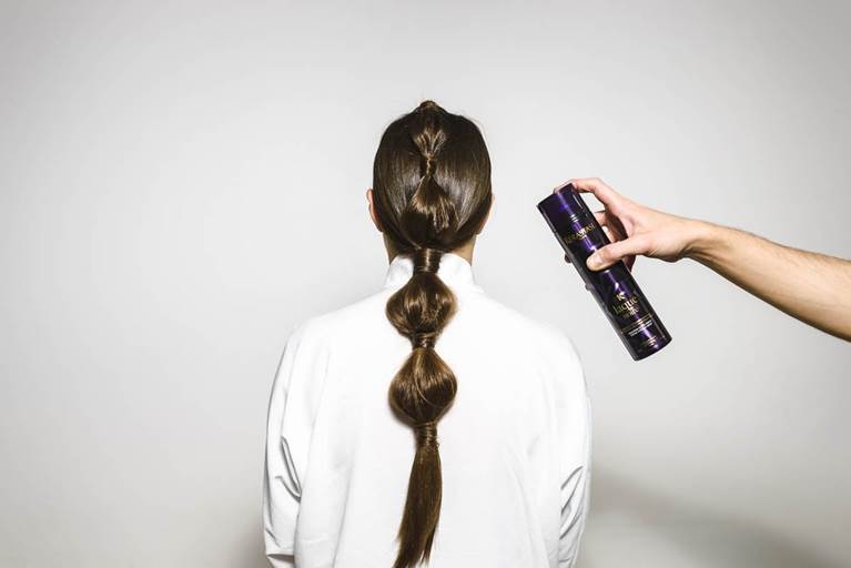 A person with a braided hairstyle facing away, while a hand holds a hairspray can to style the hair.