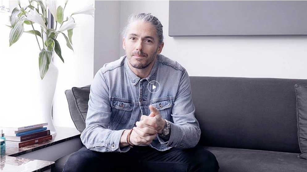 A man with long, gray hair sits on a couch, casually dressed in a denim jacket, hands clasped, in a modern living space.