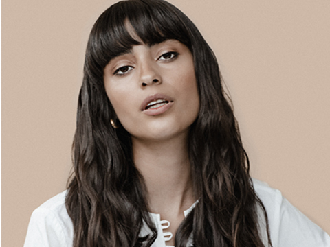 A woman with long, wavy hair and bangs, wearing a white blouse, poses against a soft beige background.