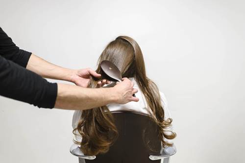 A stylist gently brushes long, flowing hair of a woman seated in a salon chair against a plain background.