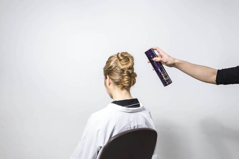 A stylist sprays hairspray onto the elegant updo of a woman, seated in a salon chair against a plain backdrop.